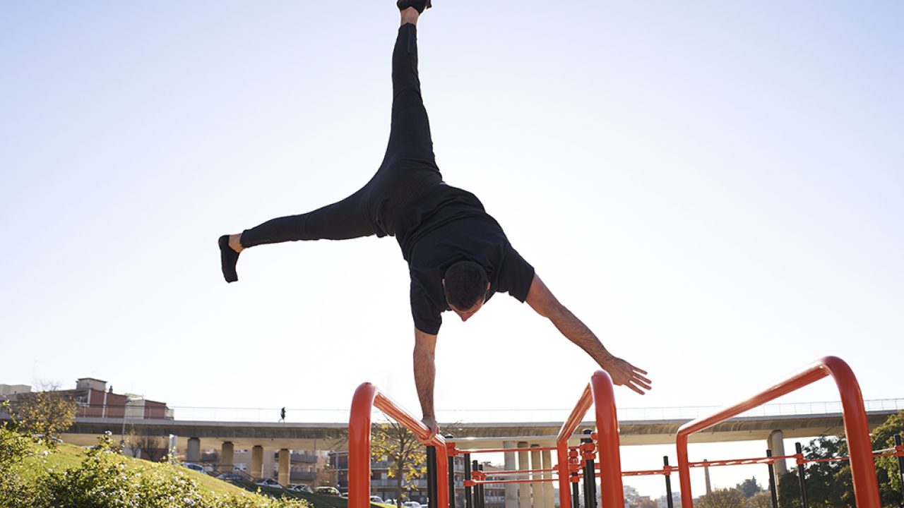 Man balancing with one hand, handstand on the bar, in a bar park in a city. Outdoor exercise. Concept of healthy living, sport, training, calisthenics.